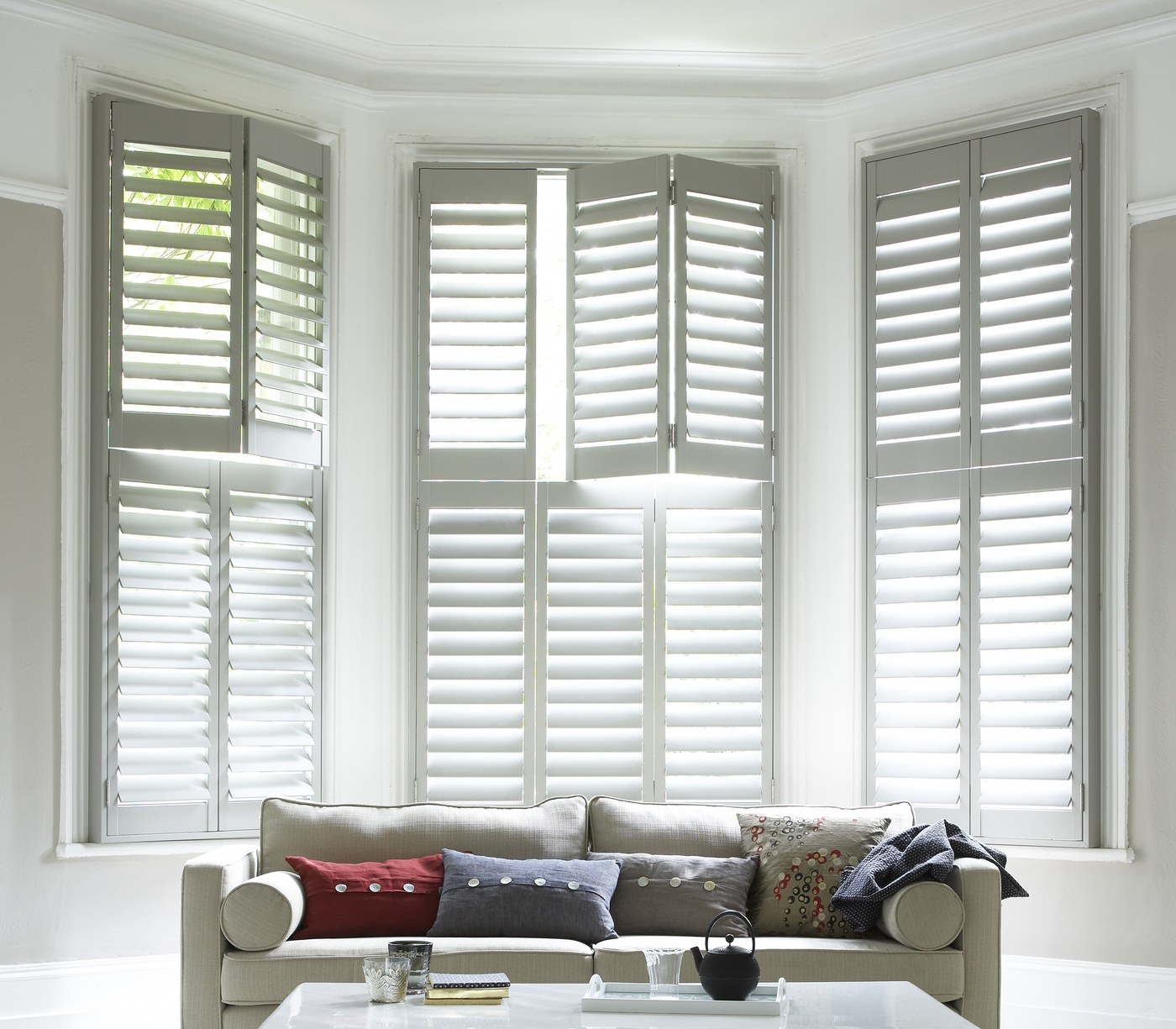 large bay window in a room dominated by white colours, with a white sofa and white large tier on tier wooden shutters