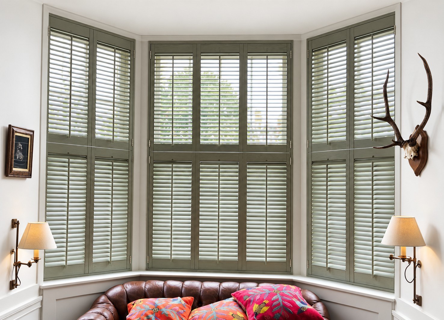 Sage green tier-on-tier shutters across a bay window above a leather sofa