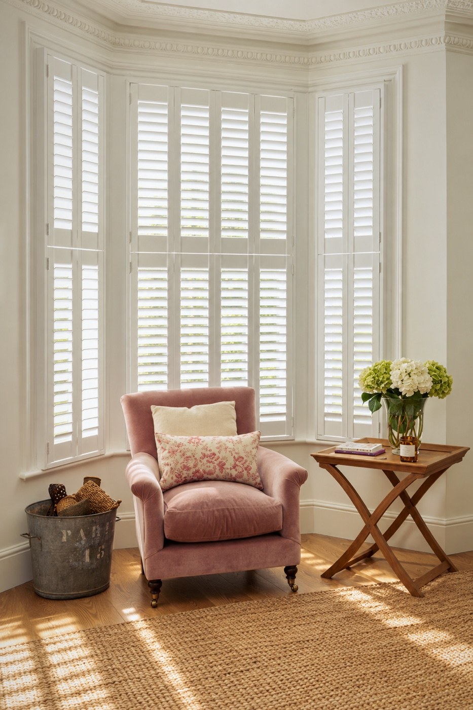 White full-height plantation shutters in a bay window, with a pink velvet armchair and hydrangeas on a side table